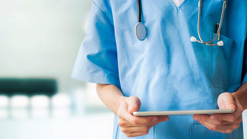 Healthcare worker in scrubs holds a tablet with a stethoscope around their neck.